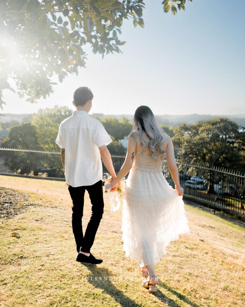 A couple walks hand in hand on a sunny day, captured beautifully by Julian Photography. The woman wears a long white dress, and the man is in a white shirt and dark pants. They stroll along a grassy slope, with trees and a fence providing the perfect backdrop for this Sydney moment.